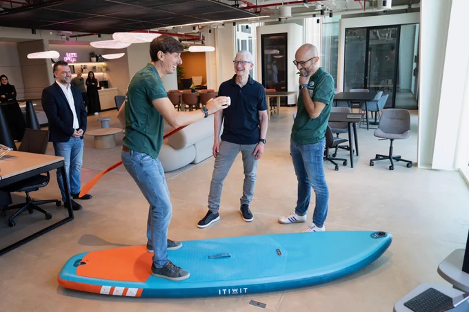 A developer from Waterspeed stands on a stand up paddleboard to demonstrate how the app works, while Tim and a few other people watch. Behind them is an office setting, with tables, chairs, and conference rooms.