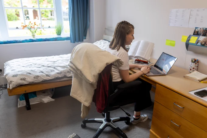 Person sitting at a laptop at a desk in a well-organized bedroom, with a bed and window in the background.