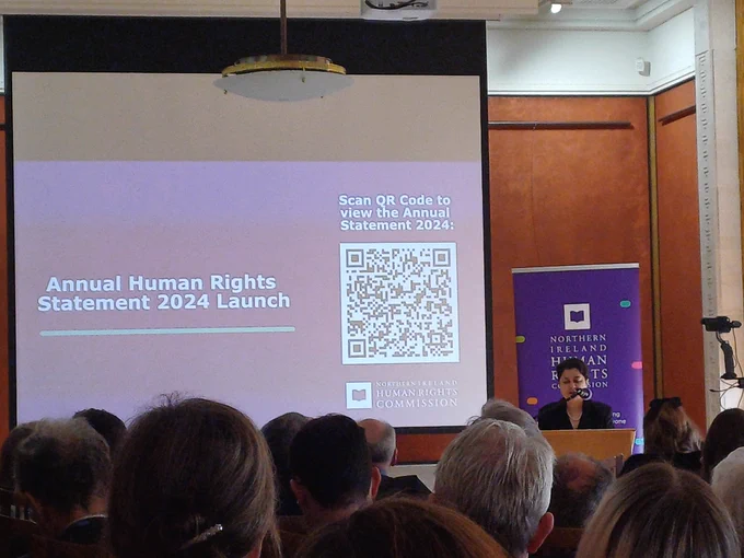 Baroness Shami Chakrabarti stands at a podium on the right of the photo, near a NI Human Rights Commission banner. A QR code is visible on a screen, leading to the annual statement.