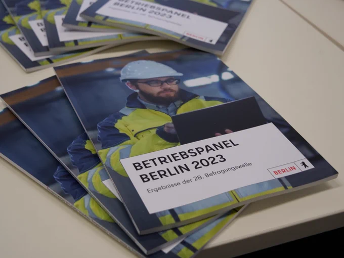 Mehrere Broschüren mit dem Titel "Betriebspanel Berlin 2023 Ergebnisse der 28. Befragung" liegen auf dem Tisch. Auf dem Cover: ein Arbeiter mit Bart und einer Brille hält ein Tablet in der Hand und trägt eine gelbe Jacke mit Reflektoren und einen weißen Bauhelm.