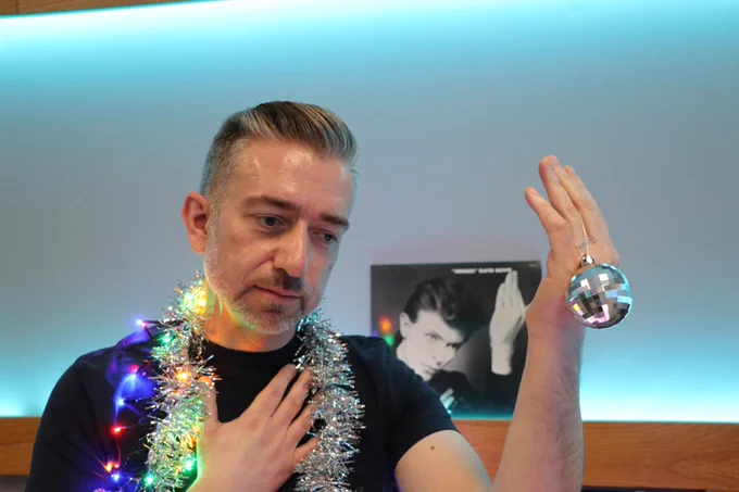 Short haired man in black t-shirt standing looking towards the ground in front of a David Bowie album recreating the look. He is wearing tinsel and fairy lights round his neck, with his right hand across his chest, and his left hand up holding a small decoration glitter ball.