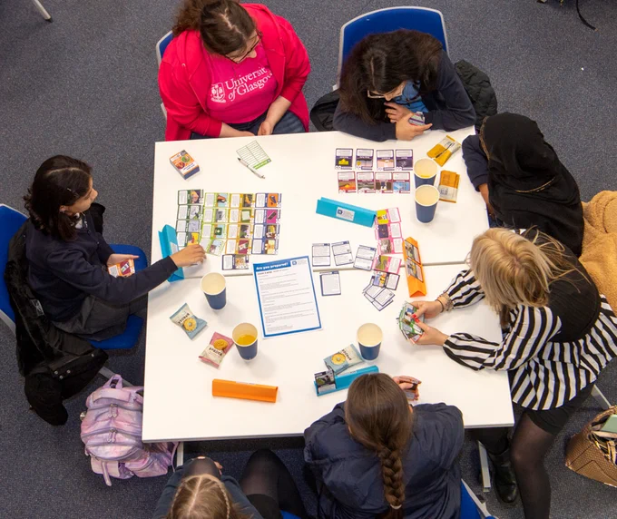 Image shows a group of people playing the card game at a table. the cards are colourful and laid out on a white table top. 