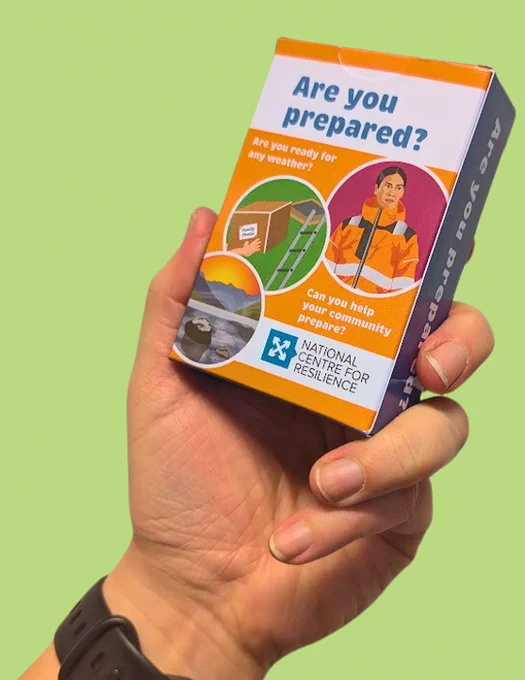 Image shows a pack of cards held up by a hand against a bright green background. The box of cards is orange and says 'Are you prepared' in blue writing on the front. The box features illustrations including of a lake and mountains, a box of family photos and a person dressed in a hi-vis jacket. 