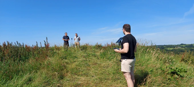 Ben Sykes and Bruce Griffith stand in the background speaking to a microphone embedded in the ground. Gerjohn in the foreground looks back to the speakers at North Wyke Farm Platform.