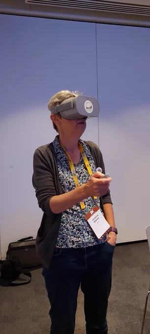 A woman wear a virtual reality headset and looks to the right of the photo. She holds the remote in her right hand, and has her left hand in her pocket.
