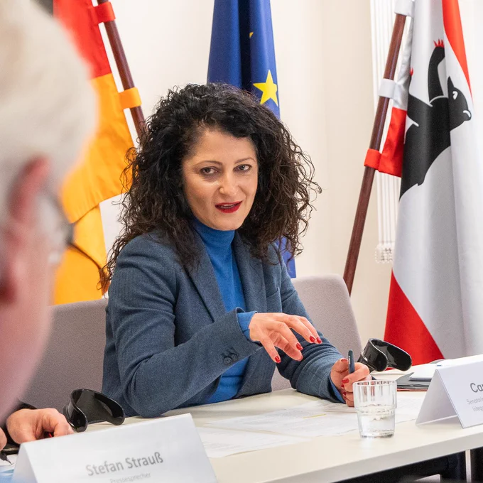 Senatorin Cansel Kiziltepe sitzt am Tisch und spricht bei der Pressekonferenz. Im Hintergrund stehen die Flaggen von Deutschlad, EU und Berlin.