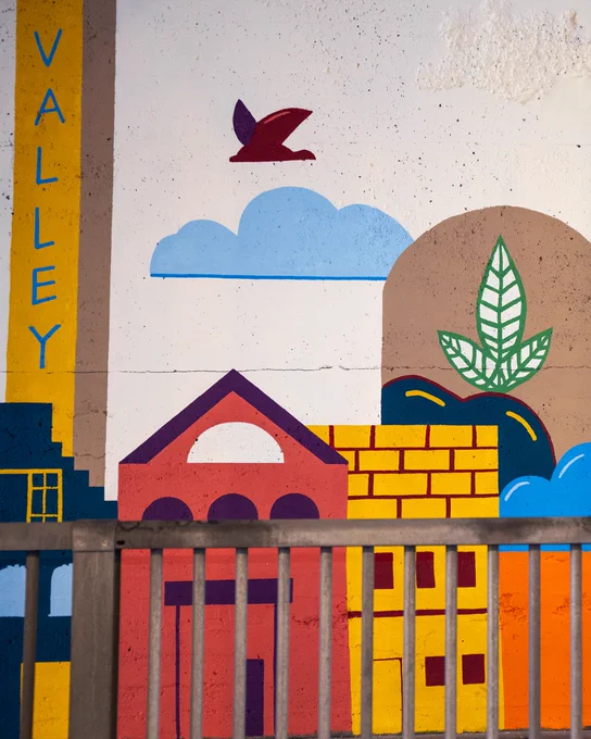 A zoomed-in panel of the mural with bold, colorful depictions of a brick building, a leaf symbol, and a bird flying above a bright yellow pillar labeled "VALLEY."

