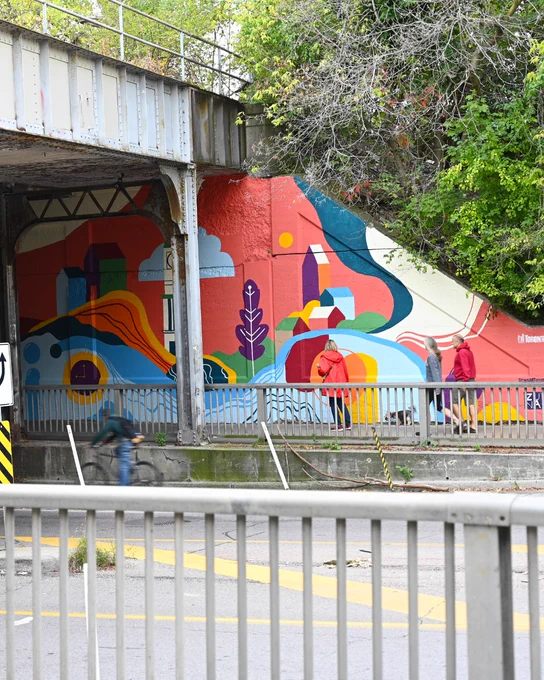  A wide-angle view of the mural at the underpass entrance. The colorful artwork includes buildings, waves, and foliage, surrounded by greenery and pedestrians walking nearby.