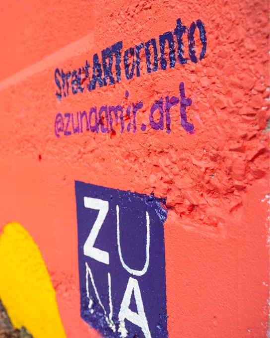 A close-up of the Scrivener Underpass Mural signature, reading "StreetARToronto @zunaamir.art" in purple text on an orange-red wall with yellow accents. Below it is a painted logo that says "ZUNA" in white letters on a purple background.
