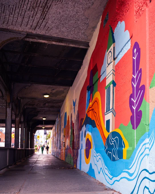 A vibrant section of the Scrivener Underpass Mural featuring abstract waves, trees, and the clock tower of Toronto’s Summerhill LCBO on a red and orange background. The perspective stretches along the underpass walkway.