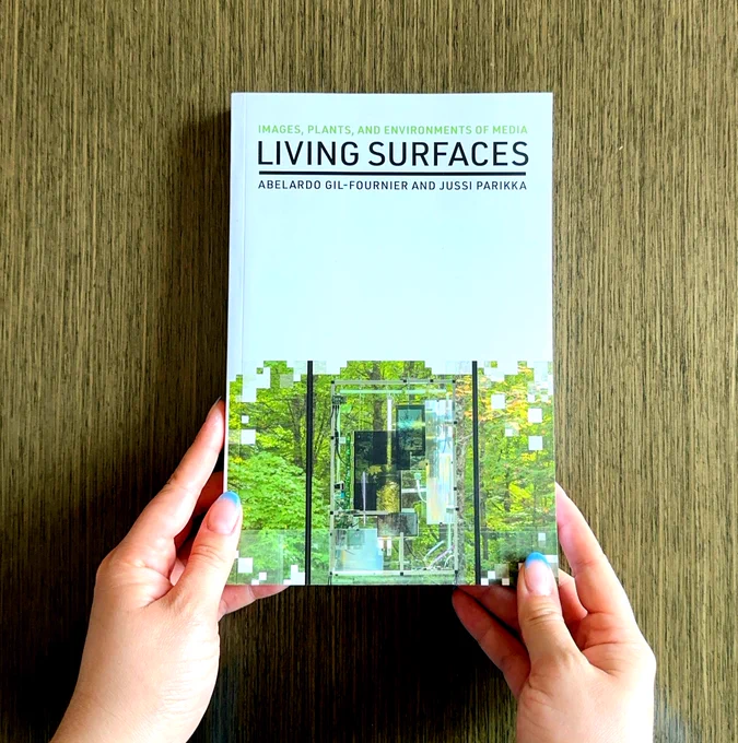 A person holding a book titled "Living Surfaces" by Abelardo Gil-Fournier and Jussi Parikka, featuring a cover design with layered images of plants and technological elements.