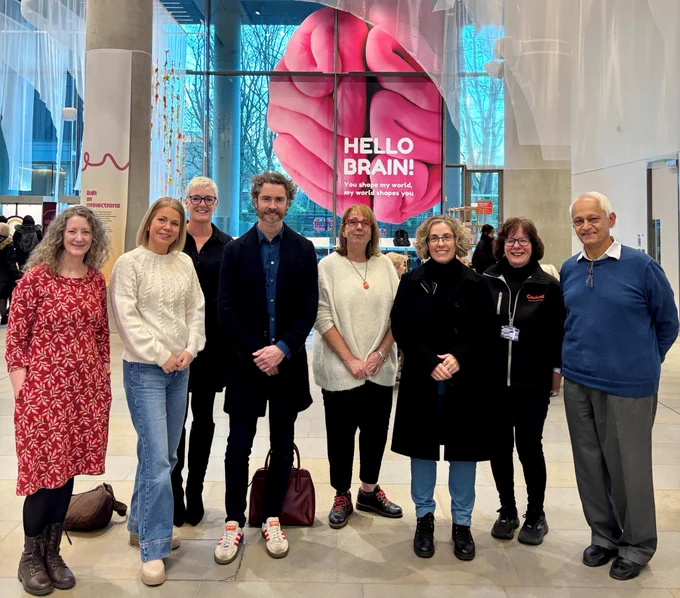 Image shows the Fifth Sense team with their guests in a large exhibition hall. Behind them is a big Hello Brain! sign and a big picture of a pink illustrated brain on a large window. The image shows left to right - Mags, Sally, Helen, Duncan, Sarah, Marianna, Mary and Azim. 