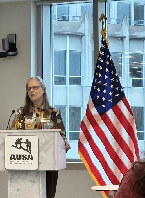 White woman with shoulder length hair standing behind grey podium next to American flag.