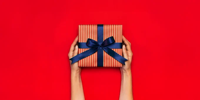 Two hands holding a striped gift box with a blue ribbon, against a vibrant red background.