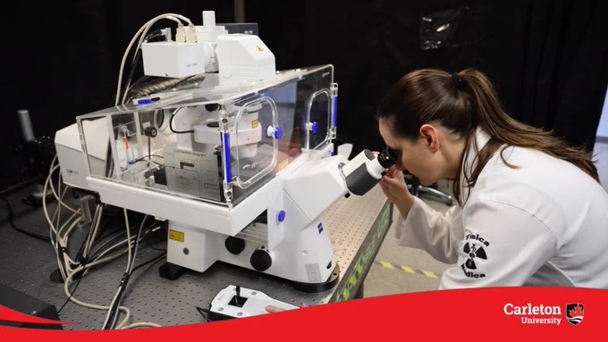 Researcher looking through a microscope in front of complex lab equipment.