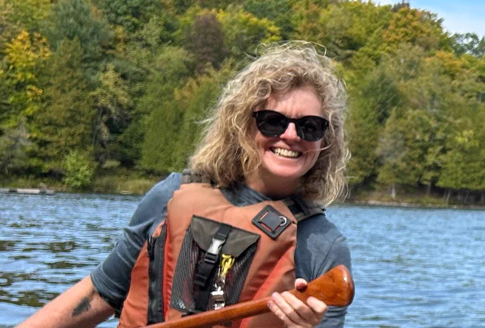 Shelley is wearing black sunglasses, a blue t-shirt and orange life jacket while paddling on a lake. 