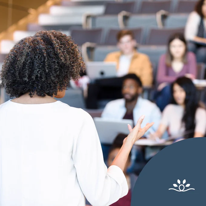 Black woman standing with her back to the camera wearing a long sleeve white shirt rolled up with shoulder length brown tightly curled hair that has lighter brown highlights. She is teaching a group of students that are blurred out to emphasize the professor practicing accessible teaching. Some students have a laptop. At the bottom left corner there is a blue semi circle with a national disability logo of a icon with a thick white line that loops in a circle in the middle and curves down and upward at each end. Above the line are four white spear-shaped leaves arranged in an arc over the loop.
