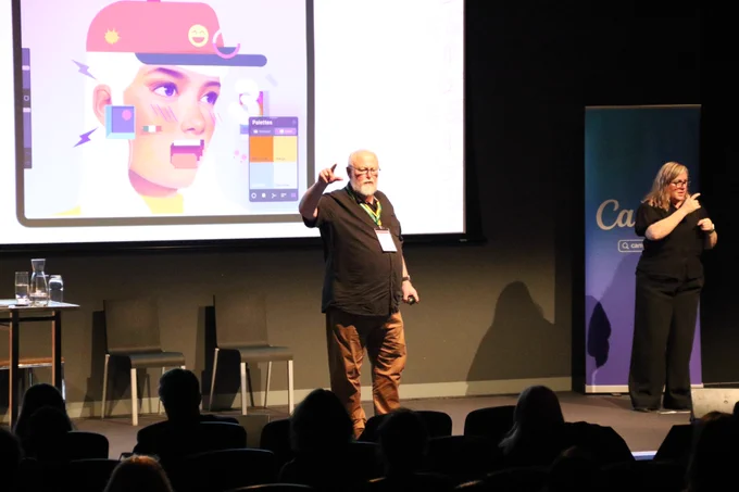 Greg Alchin pointing out towards the audience while presenting on stage at  A11y Camp. Behind him on the screen is an in progress digital drawing of someone wearing a cap, showing a colour palette. On the right of stage is an Auslan interpreter.