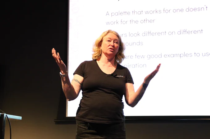 Donna Spencer gesturing with her arms open on stage at A11y Camp, behind her is some text on a widescreen from her presentation slide.