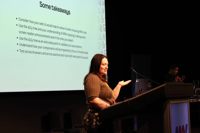 Sarah Federman presenting at the lectern at A11y Camp, gesturing to the slide behind her on the big screen.