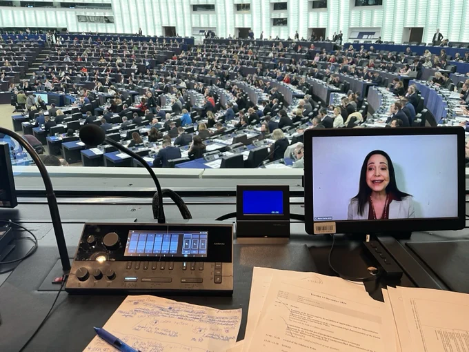 A view from an interpreter's booth into a full Hemicycle Chamber of the European Parliament. In the foreground, notes and documents are seen next to the interpreter's console and microphone as well as a video screen showing Ms María Corina Machado remotely delivering her speech.