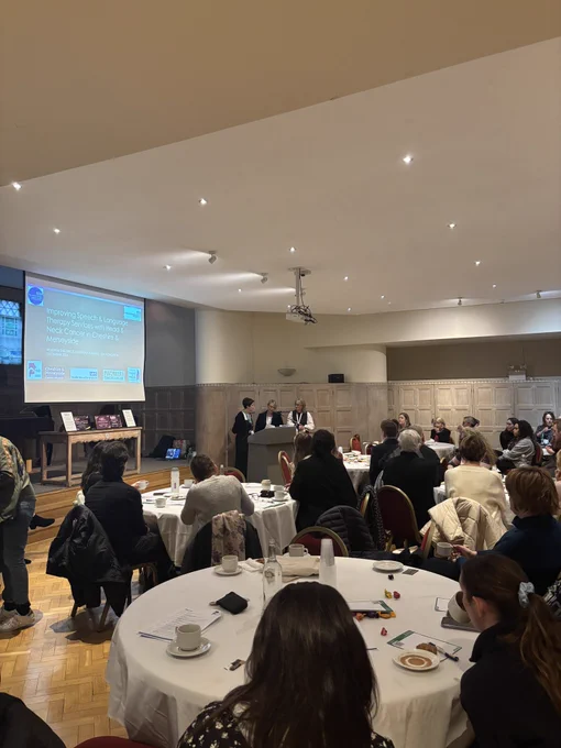 A room of people sitting at round tables listen to three women who are presenting. There is a visual on a screen introducing Heulwen, Lisa and Catriona.