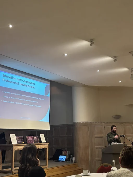Josh is standing at a podium speaking to the room. He has a dark beard and is wearing a green shirt. There is a screen reading Education and Continuous Professional Development.