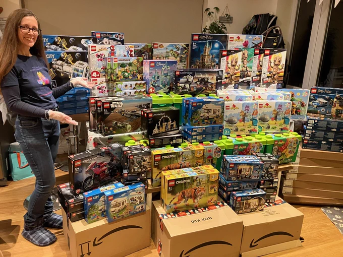 Ginny standing in a room surrounded by numerous LEGO sets, gesturing towards the collection with a smile. The sets include various themes like LEGO City and LEGO Technic.