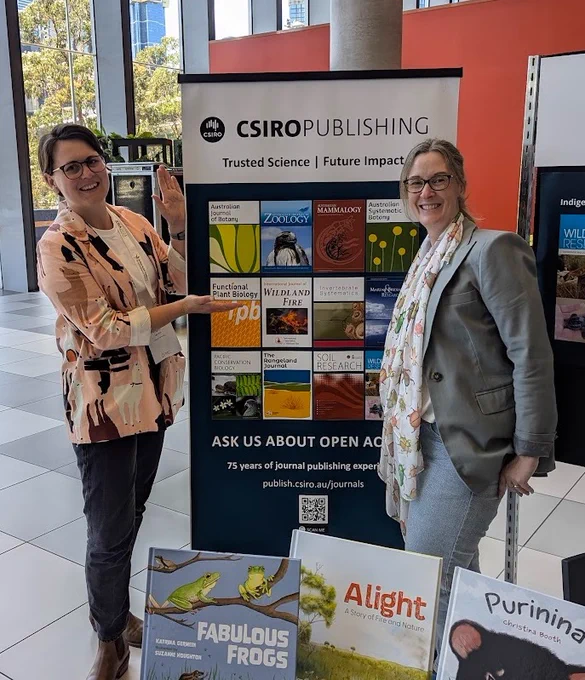 Two people are standing either side of a banner, with childrens books in the foreground. The banner features the cover art of twelve different scientific journals, including the Australian Journal of Botany.