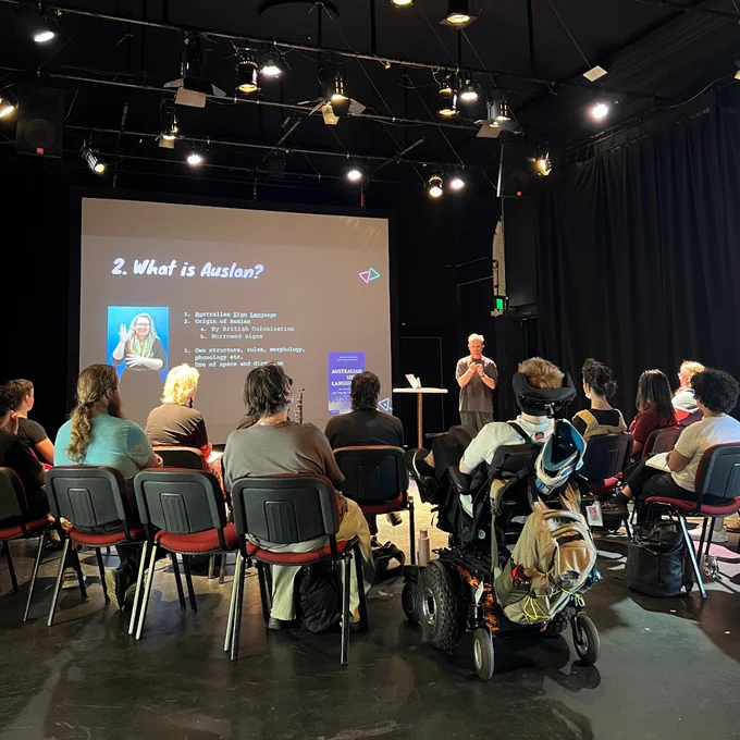 A group of people watching artist Luke D King giving a presentation. There is a large screen in the background showing a slide with the heading 'What is Auslan'. 