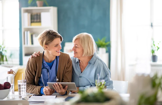 A nurse sits together with an older patient in a home setting. They are sat side by side smiling; the patient has her arm around the nurse's shoulder.