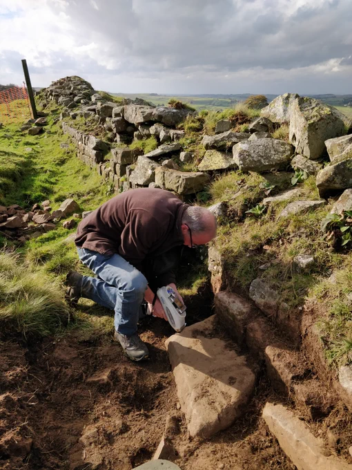 A geologist using a portable xray fluorescence (pXRF_ to analyse the chemical composition of Hadrian's Wall stones and Whin Sill bedrock. The line of Hadrian's Wall stretches along behind him. 