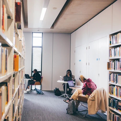 Three students sit at seperate tables with open laptops in front of them. On either side of them are shelves of library books.