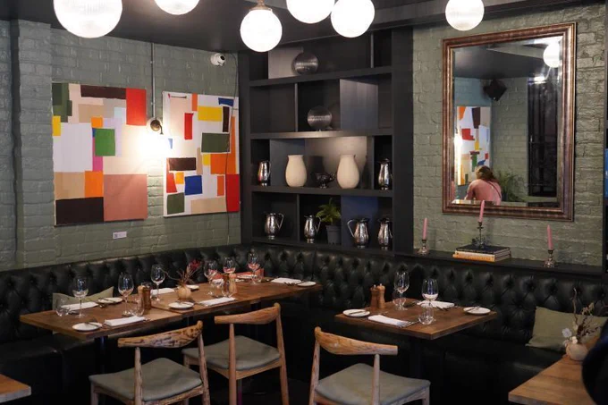 A restaurant interior featuring a green-painted brick wall adorned with colourful abstract paintings. The space includes polished wooden tables set with glassware and plates, and rustic wooden chairs. A large mirror with a wooden frame and decorative shelves display vases, pitchers, and small plants.