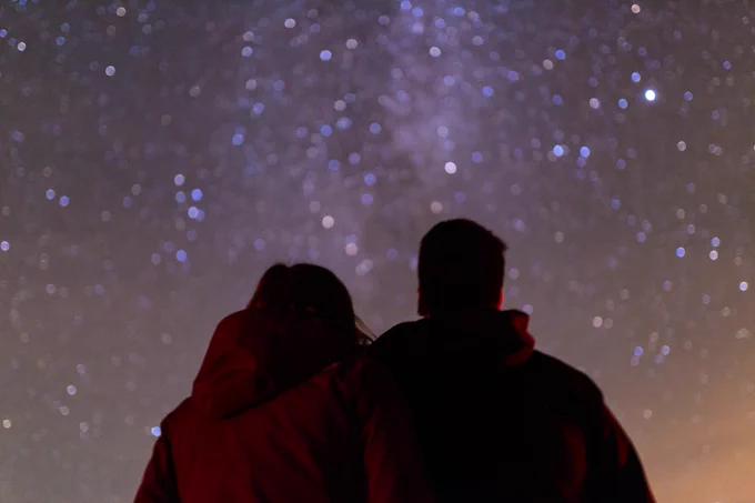 There is a woman and man standing with their backs to the camera. The woman on the left is wearing a red jacket, the man on the right is wearing a black puffer jacket. There is a slightly blurred image of a starry night sky above them.