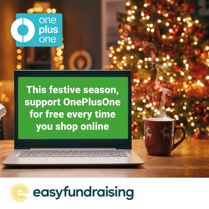 Image shows a laptop in the foreground with the words, 'This festive season, support OnePlusOne for free every time you shop online,' on the screen. There is a Christmas mug on the table next to the laptop and a blurred lit Christmas tree in the background. The OnePlusOne logo appears in the top left-hand corner.