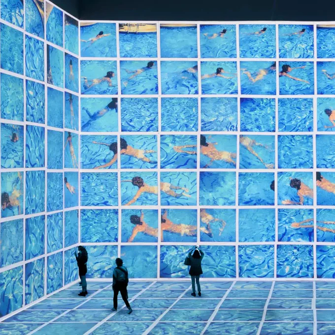 People in exhibition space looking at exhibition by David Hockney. Imagery is of blue swimming pool artwork by Hockney with multiple people swimming. Artwork is divided up into squares.