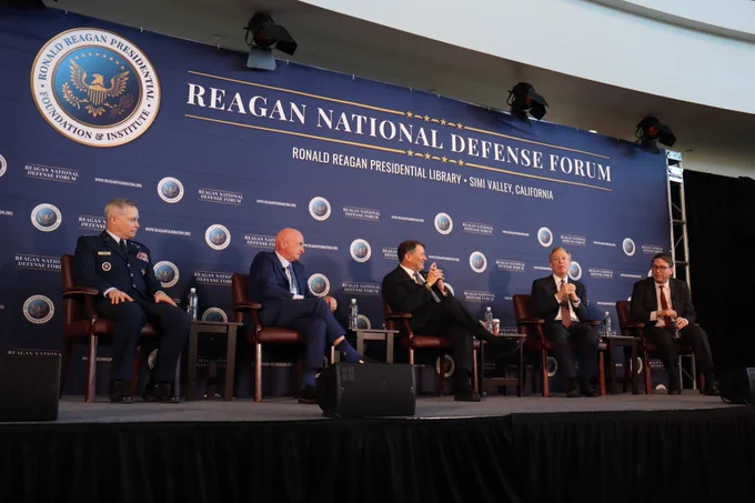 A panel of men sit on a stage in front of a sign that reads, "Reagan National Defense Forum."