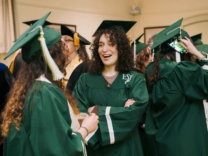 Graduate in green cap and gown smiling joyfully while conversing with classmates at a graduation ceremony of the State University of New York College of Environmental Science and Forestry (SUNY ESF).