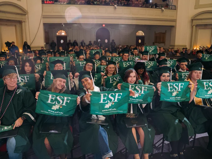 Graduates seated in a ceremonial hall holding up banners with "ESF" representing State University of New York College of Environmental Science and Forestry, during a graduation ceremony.