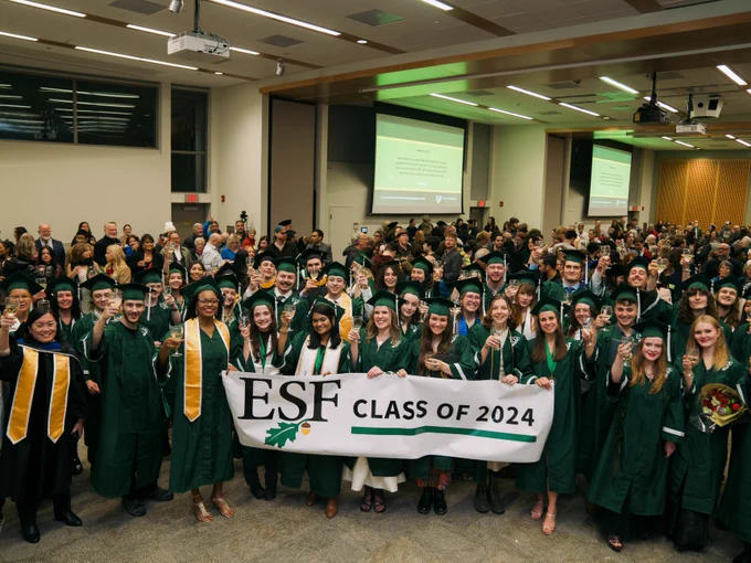 Graduates holding a 'ESF Class of 2024' banner at a commencement ceremony, with attendees in the background.