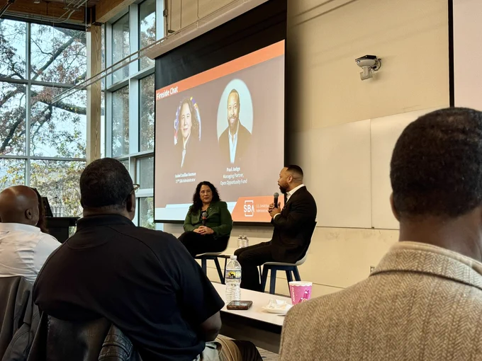 Two individuals sit in chairs having a discussion at a panel in front of a screen displaying a presentation titled "Fireside Chat" with photos and names of two featured guest speakers, Administrator Guzman and Paul Judge. Audience members are seen from behind, listening attentively in a room with large windows