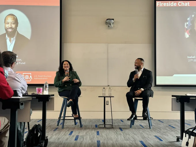 Two individuals seated in chairs on a stage in a lecture hall, engaged in conversation during a 'Fireside Chat' event. The background includes a projection of the event title and names of the participants, Isabel Guzman from the U.S. Small Business Administration. Audience members are visible in the foreground.