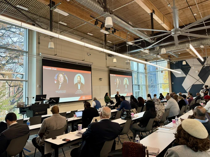 Audience members seated at a conference with a presentation featuring two speakers on a large screen in a hall.