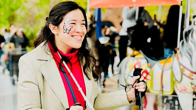 Xian has a butterfly painted on her cheek. She is wearing a red shirt with a tan coat and smiling as she holds her ski poles while near a carousel 
