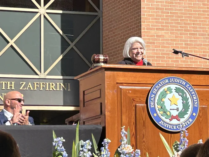 Senator Judith Zaffirini wearing a red sweater and dark jacket smiles as she stands at a large brown wooden lectern in front of the newly named Dean Senator Judith Zaffirini Justice Center in Laredo, Texas.