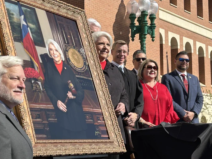 Officials stand around large gold-framed portrait of Dean Senator Judith Zaffirini holding gavel with Texas flag in background.