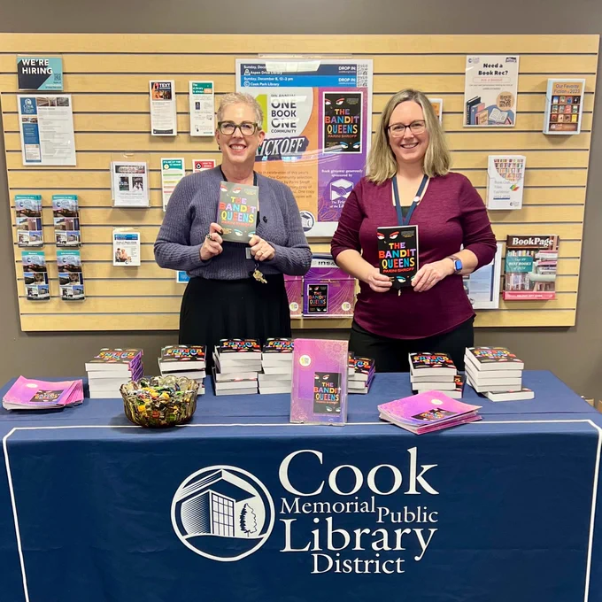 Two staff members pose in front of the library table showing off the One Book, One Community book The Bandit Queens by Parini Shroff.