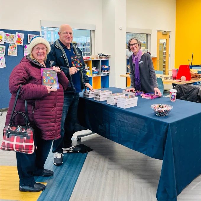 Three people pose in front of the library table showing off the One Book, One Community book The Bandit Queens by Parini Shroff.