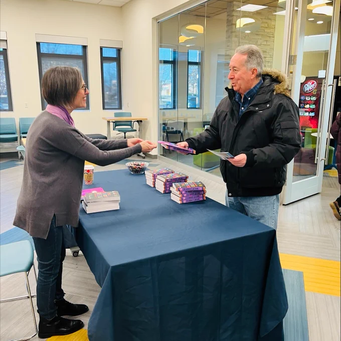 A staff member hands a patron a free copy of the One Book, One Community book The Bandit Queens by Parini Shroff.